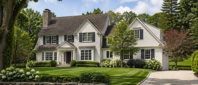 A classic white house with black shutters and a gabled roof sits among lush greenery and trees in New Canaan