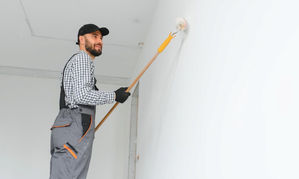 Painter in workwear rolling white paint onto a wall using a long-handled roller.