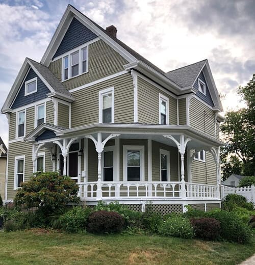 A charming Victorian-style house with olive green siding and white trim features a wraparound porch and detailed woodwork.
