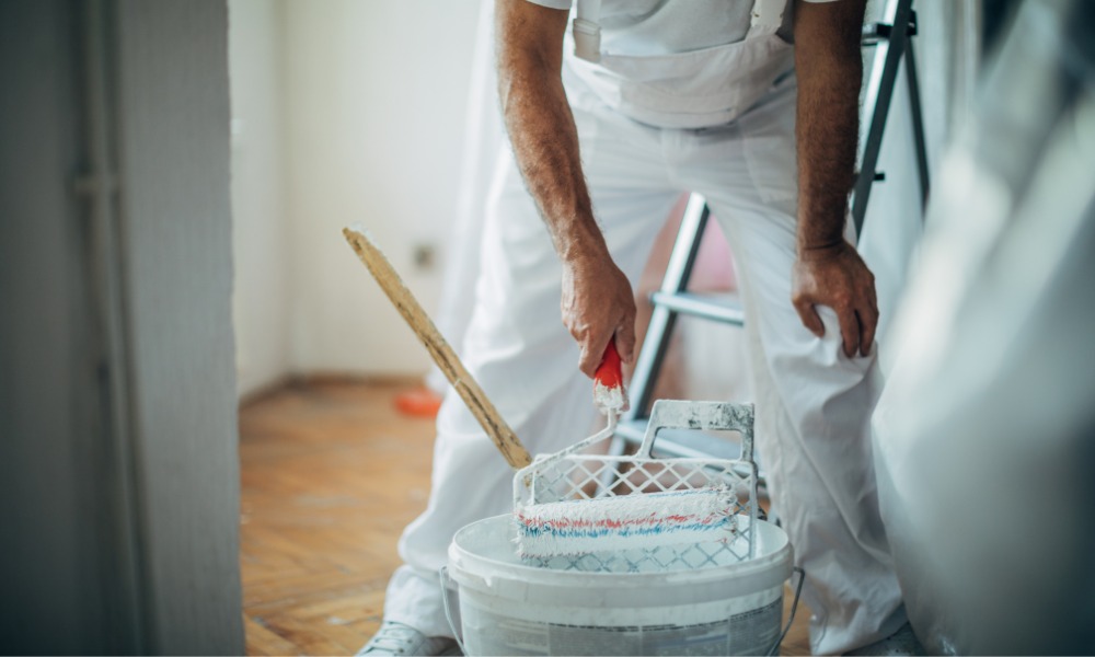 A painter dips a roller into a white paint bucket