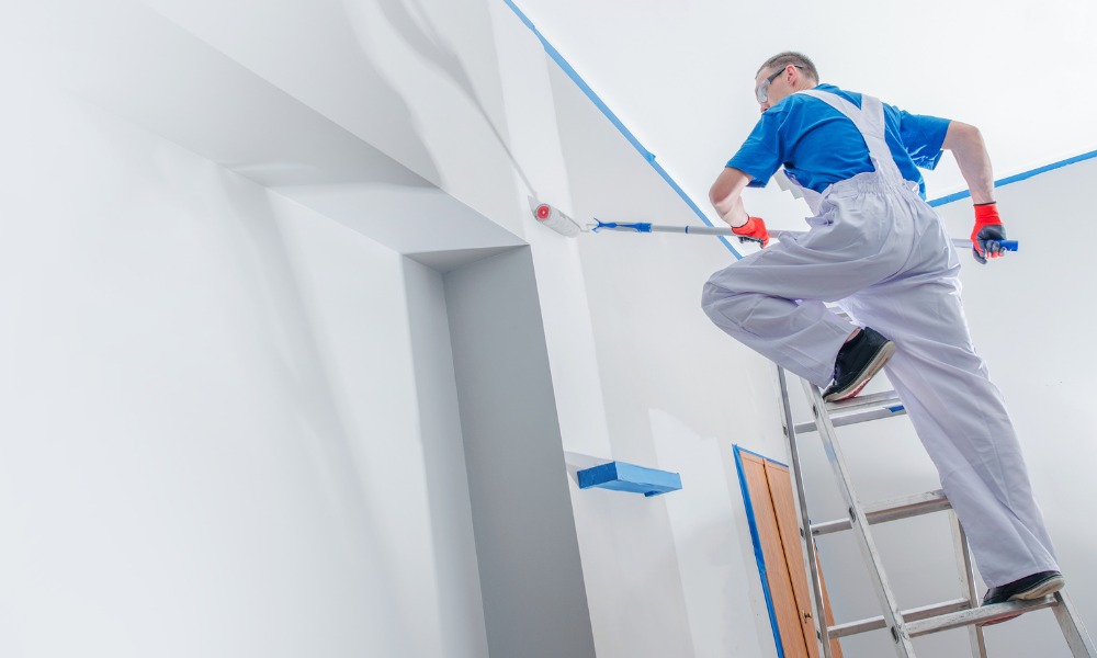 A painter on a ladder uses a roller extension pole to paint a ceiling
