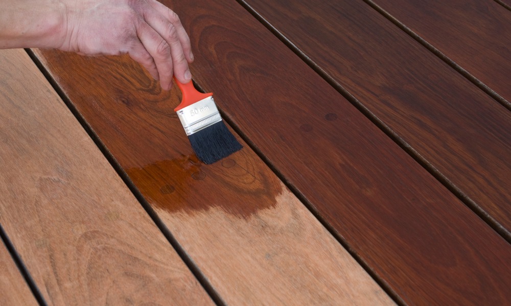 Hand using a paintbrush with an orange handle to apply dark wood stain