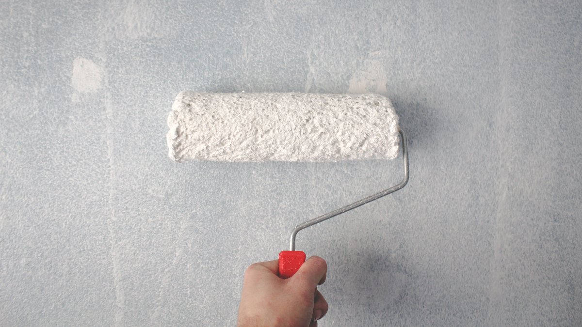 Person applying white paint on a wall with a roller in an indoor setting.